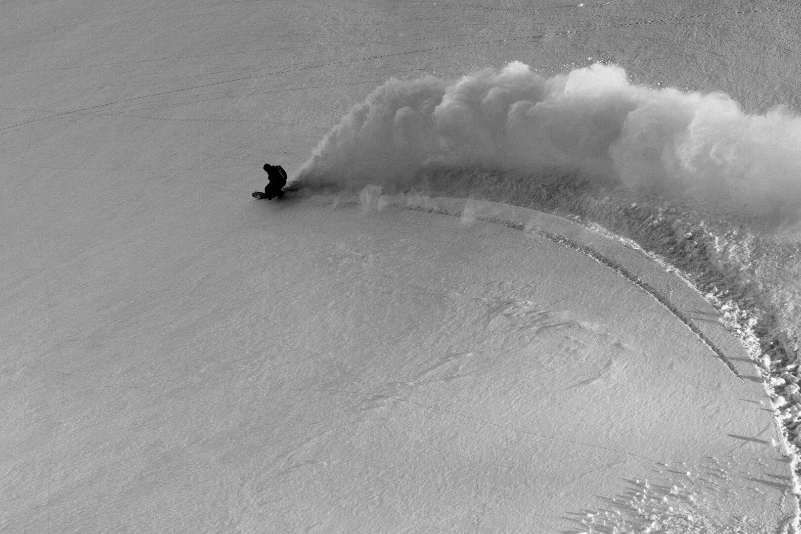 A snowboarder trailing a long plume of dry powder across an open face